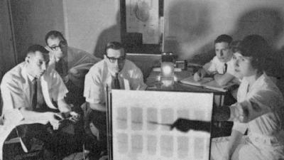 old photo of group of four men and one woman looking at a screen