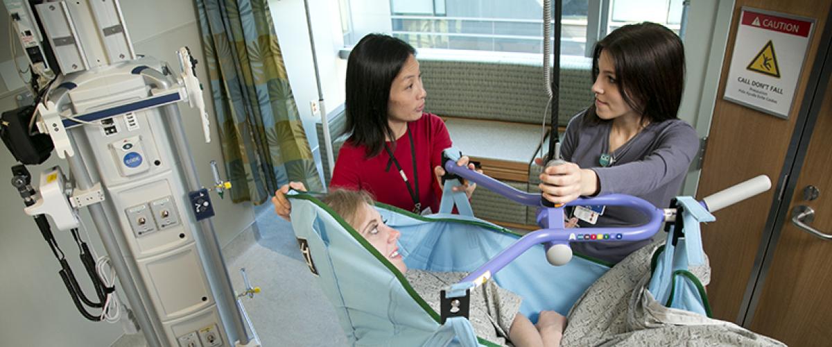 two women help lift a patient in lift equipment in a hospital room